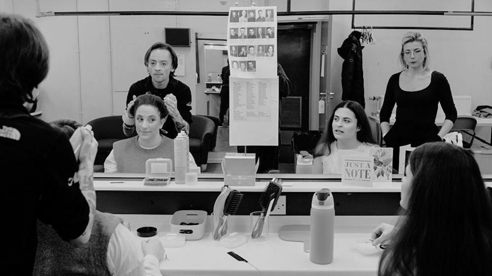 Emily Webb and Rebecca Gibbs sit at a dressing table facing the mirror with two stylists stood behind them