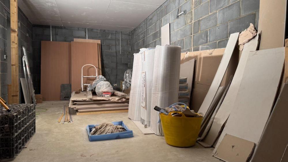 A room, finished in bare blockwork, is filled with plaster board, insulation, doors and a step ladder, with buckets of debris left.
