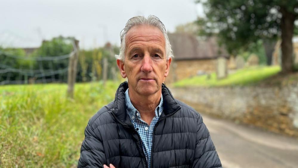 Nick Frampton with short white hair wearing a black raincoat and standing on grass in front of a church