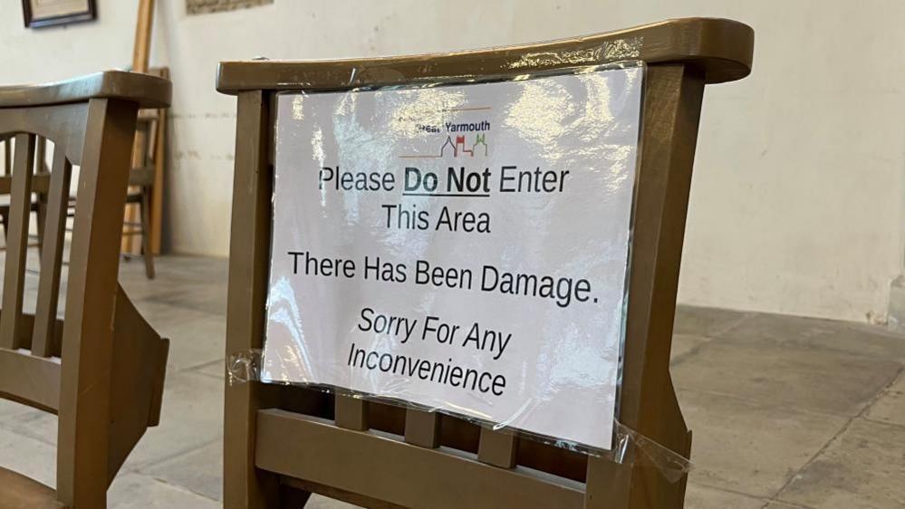 A sign, laminated in plastic, giving off a relection, is taped to a chair, forming a corden. It states: "Great Yarmouth Minster. Please do not enter this area. There has been damage. Sorry for any inconvenience."