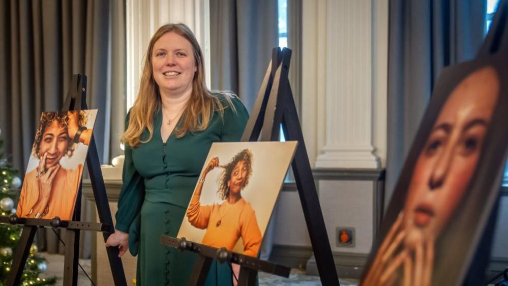 A woman in a long bottle green dress stands between photographs which are being displayed on black easels. 