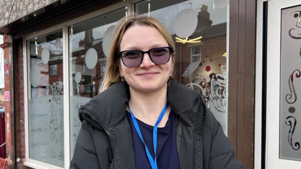 Florentine Apetrel with long light brown hair and large tinted glasses, smiling at the camera. She is wearing a large black raincoat and a blue shirt with a blue lanyard. She is standing next to a shop.