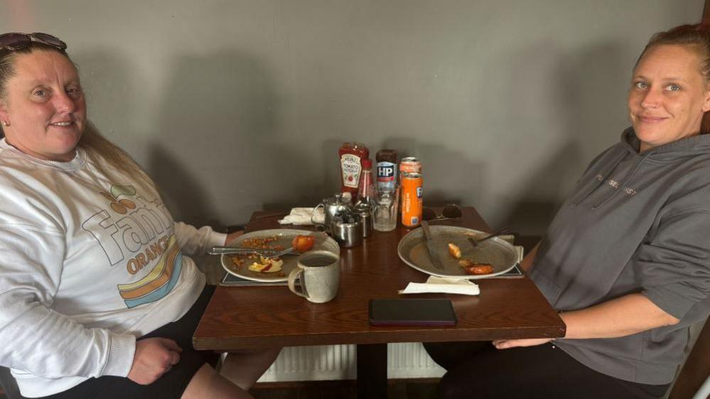 Vicky dressed in a white shirt sits at a café table opposite her friend Mandy. They have plates of leftover food in front of them.