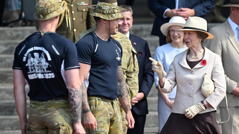 Princess Anne on the right of the picture, wearing a cream hat and jacket, talking to service personnel wearing fatigues