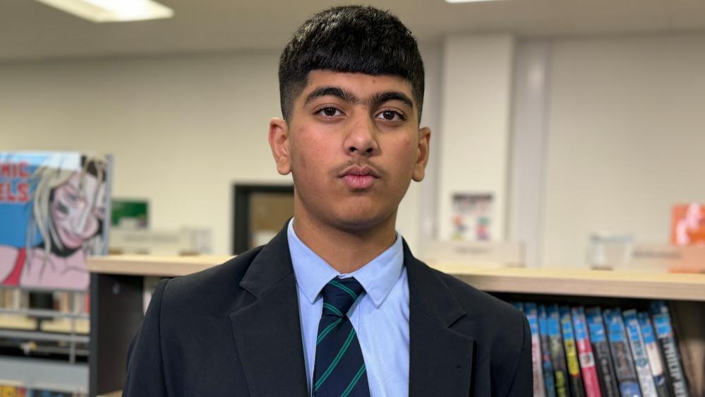 A head-shot a school boy in a blue shirt and navy blazer. He is looking straight into the camera and has short black hair with a short fringe. There is a bookcase behind him.