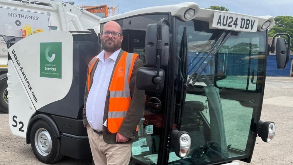 Councillor Paul Wells, wearing a white shirt, brown jacket and orange high-viz vest with reflective silver strips, and cream coloured chinos, is pictured next to a Great Yamrouth Borough Services road sweeping machine, within the depot with buildings and a dustcart in the background.