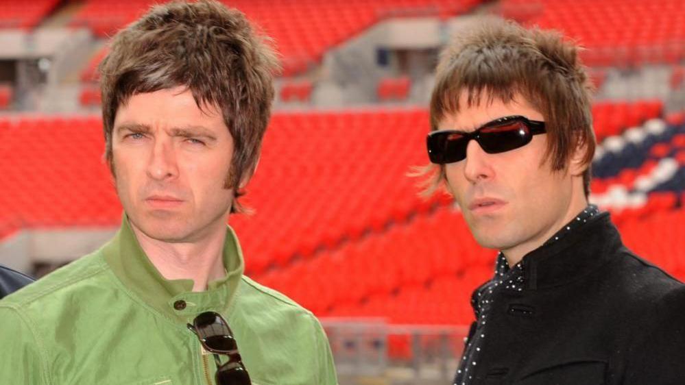 Noel Gallagher pictured on the left, in a light green jacket, and his brother, Liam, wearing shades and standing to Noel's right, wearing a black jacket and black and white cravat.