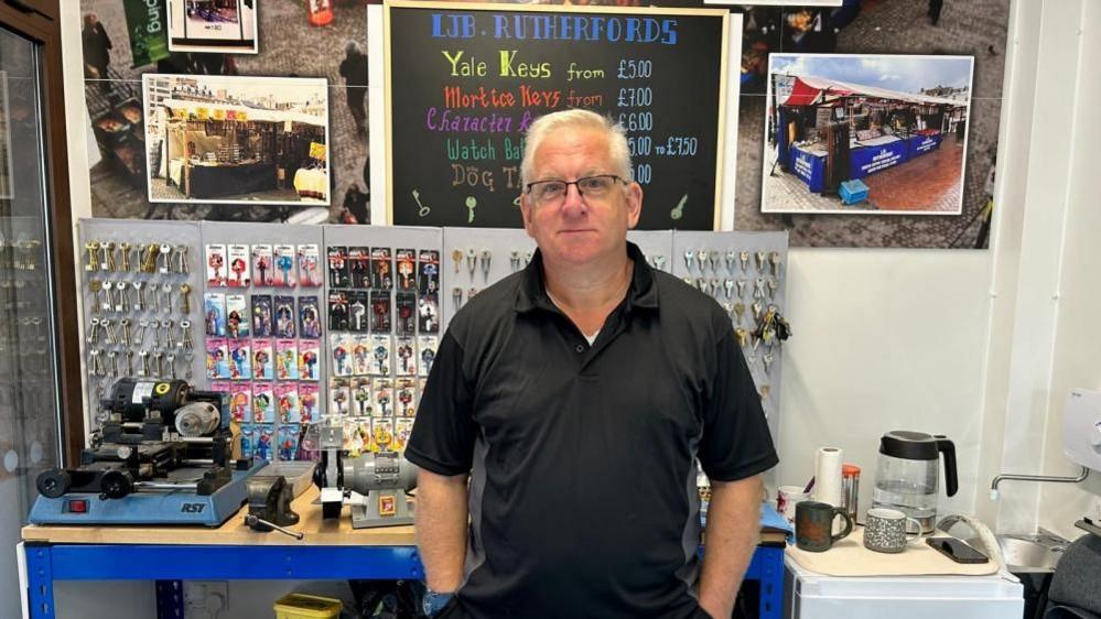 Les Brennan with short light-coloured hair and glasses, wearing a black T-shirt with his hands in his pockets.  Various keys are on display on a board behind him.