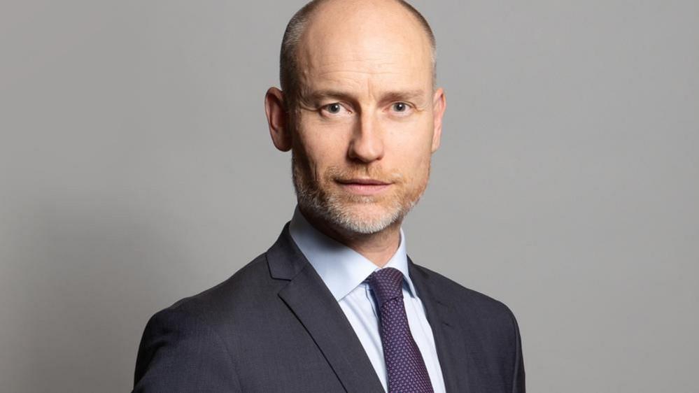 Stephen Kinnock: a bearded man, with tight cropped brown and grey beard wearing pale shirt, blue and red tie and grey jacket  