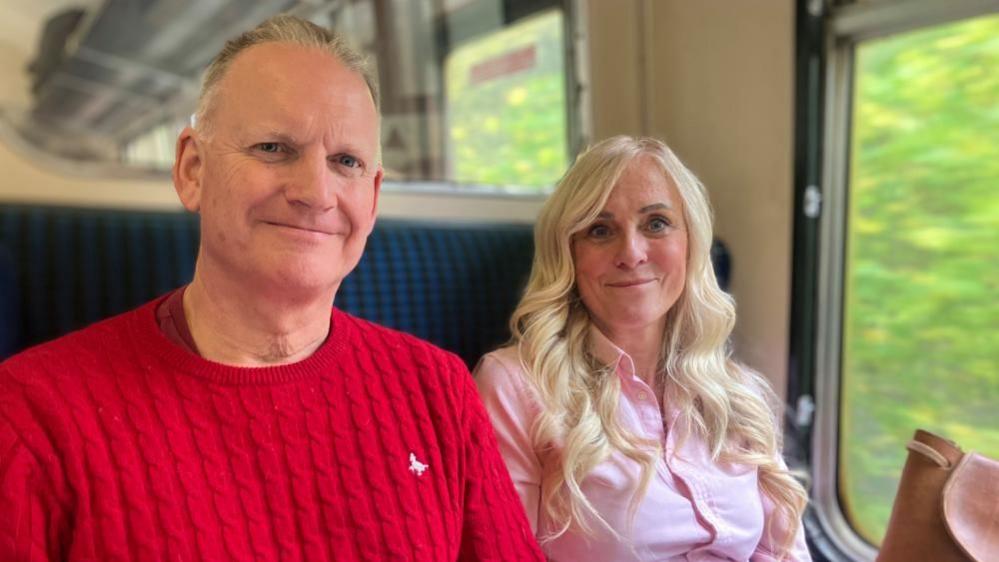 A white man and woman sit happily together in a heritage railway carriage while a view of green trees can be seen whizzing by through the window. The woman has long, blond hair and wears a pink shirt. The man has short, grey hair and wears a red jumper.