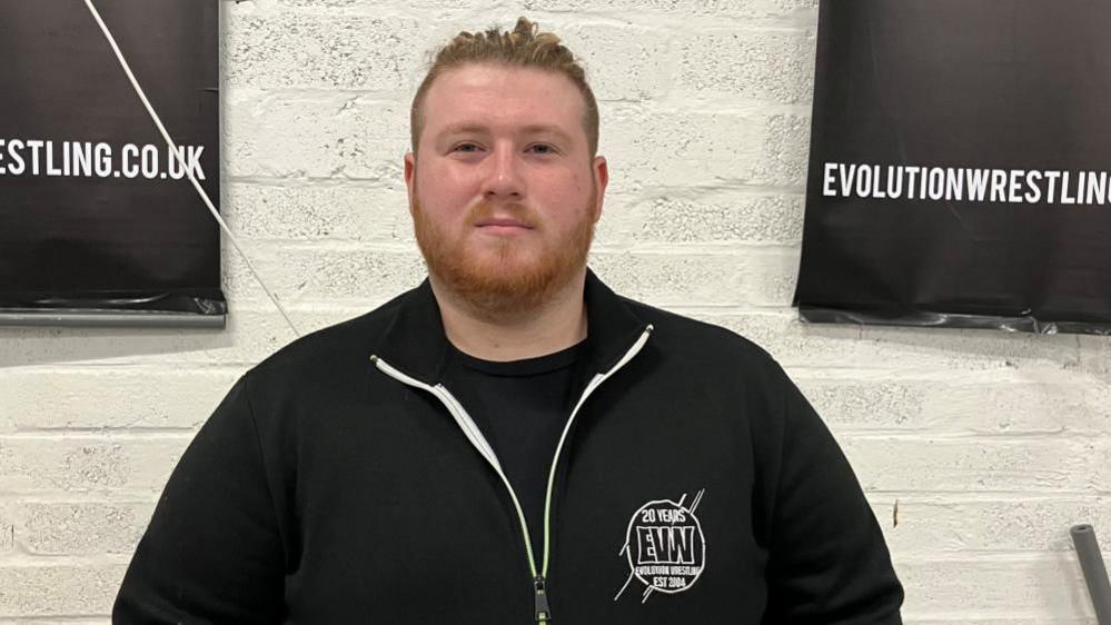 Photo of Liam Robinson, head coach at Evolution Pro Wrestling. He is wearing a black top and has a beard with his hair tied in a bun.