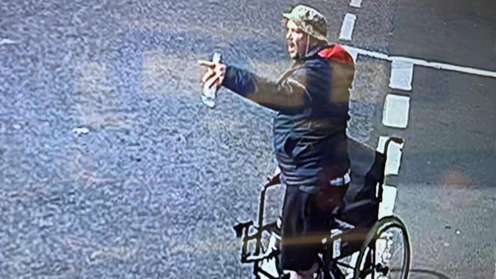 Michael McQueen standing up near his wheelchair, which is stood on the white lines of the junction of St Peter's Road with King Street in Great Yarmouth. He is wearing black trousers, and a blue jacket, which has a red hood lining. He is also wearing a hat, and holding a bottle of beer in his left hand.