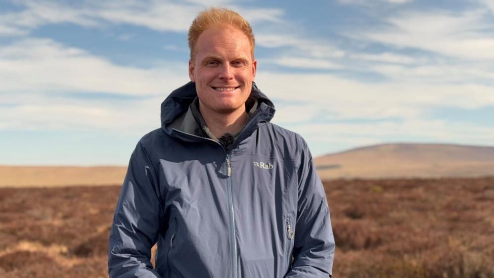 Dr Fewster is standing on a peatland smiling at the camera. He has ginger hair and is wearing a navy waterproof Rab coat. In the background are some hills.