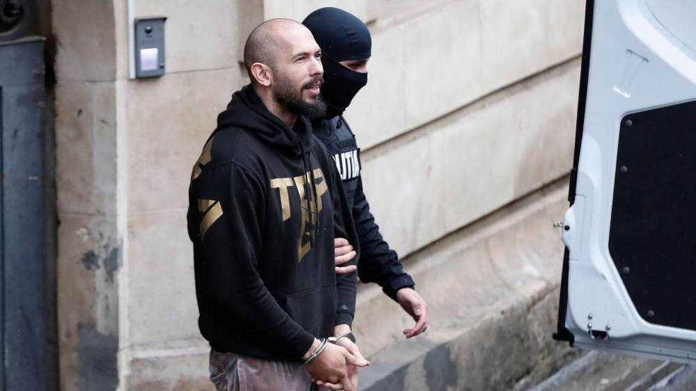 Andrew Tate (L), accompanied by a masked police officer, leaves the building after a hearing on a British arrest warrant, held at the Bucharest Appeal Court, in Bucharest, Romania, 12 March 2024