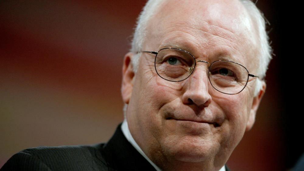 Dick Cheney wears a dark suit and white shirt while looking to his right while standing at a podium.