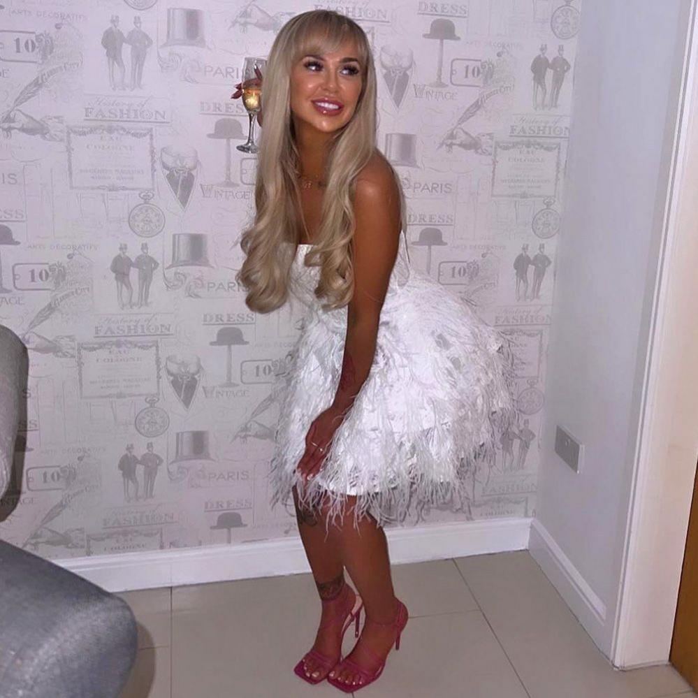 Louise - a young woman with long straight blond hair - posing at home in a white party dress and pink high-heeled shoes. She is holding a glass of white wine and standing next to a wall with grey patterned wallpaper.