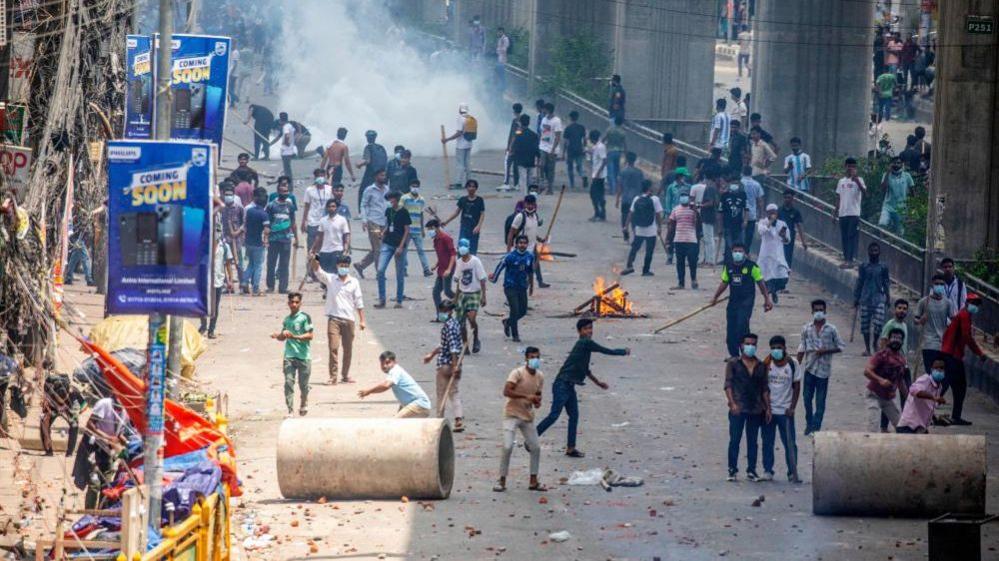 Protesters throw sticks and stones from behind concrete pipes being used as barricades. smoke billows in the background.