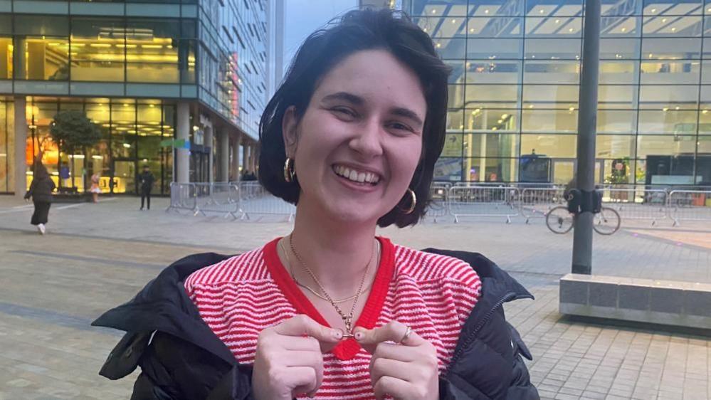 A young woman with short dark hair stands outside holding up a gold fob chain which is around her neck