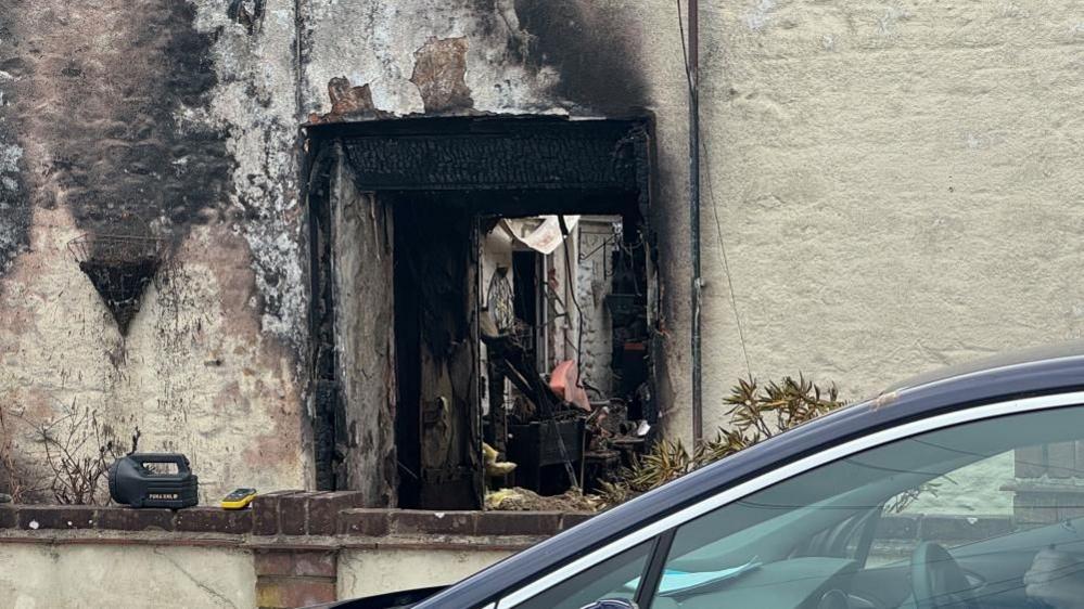 A view through the burnt-out door of the house, devastated by fire. The view shows through the house to the backyard. Smoke and fire damage has stained the walls outside. There is a car parked in the foreground with blue paint, chrome trim and glass on show.