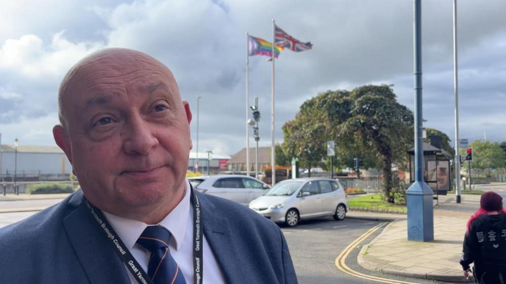 Carl Smith, dressed in a blue suit and looking to the right of the camera, stands in a car park with the Progress Pride flag and Union Jack flag flying on two masts behind him. 