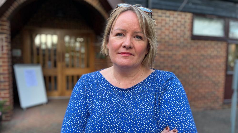 Erica Fletcher, standing outside a church in Bedford. You can see a blurred brick building with double wooden doors behind her She is looking straight at the camera, with a concerned look on her face, with chin= length blond straight hair. She has a pair of glasses on her head and is hearing a blue and white spotted top. Her arms are crossed across her chest.