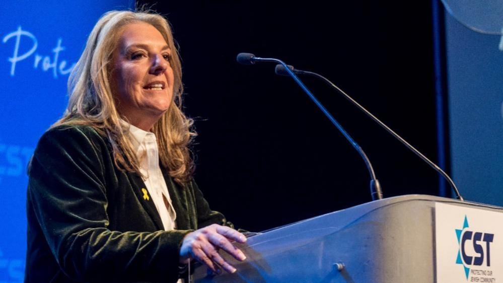 A woman with long blonde hair wears a green velvet blazer and stands speaking at a podium. 