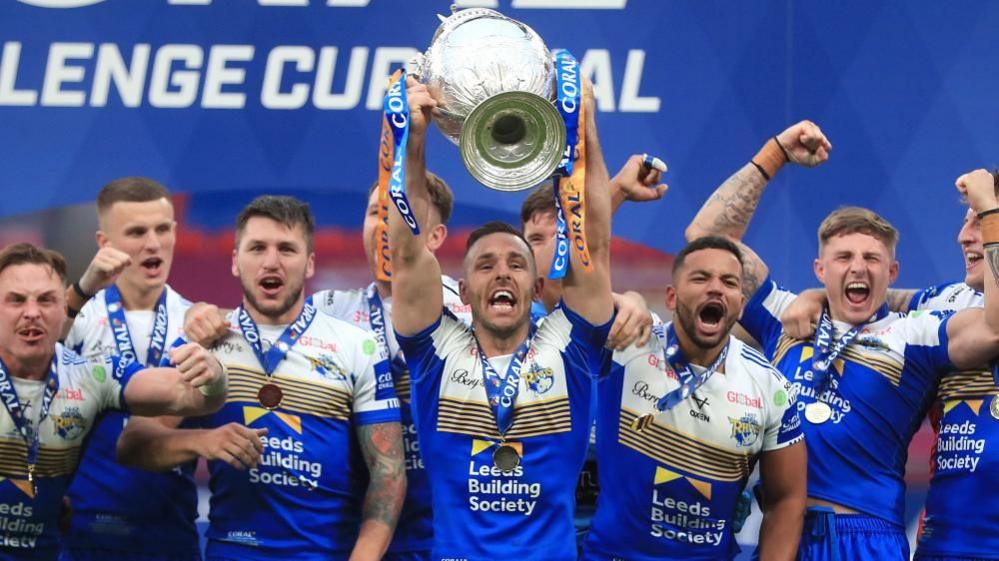 Leeds Rhinos players surround their team mate, Luke Gale as he lifts the large silver Challenge Cup trophy high above his head. The cup is adorned with blue and gold ribbons. the same colours as the Rhinos kit worn by the team. The players, who all have medals around their necks, raise their arms in jubilation as they cheer into the camera.