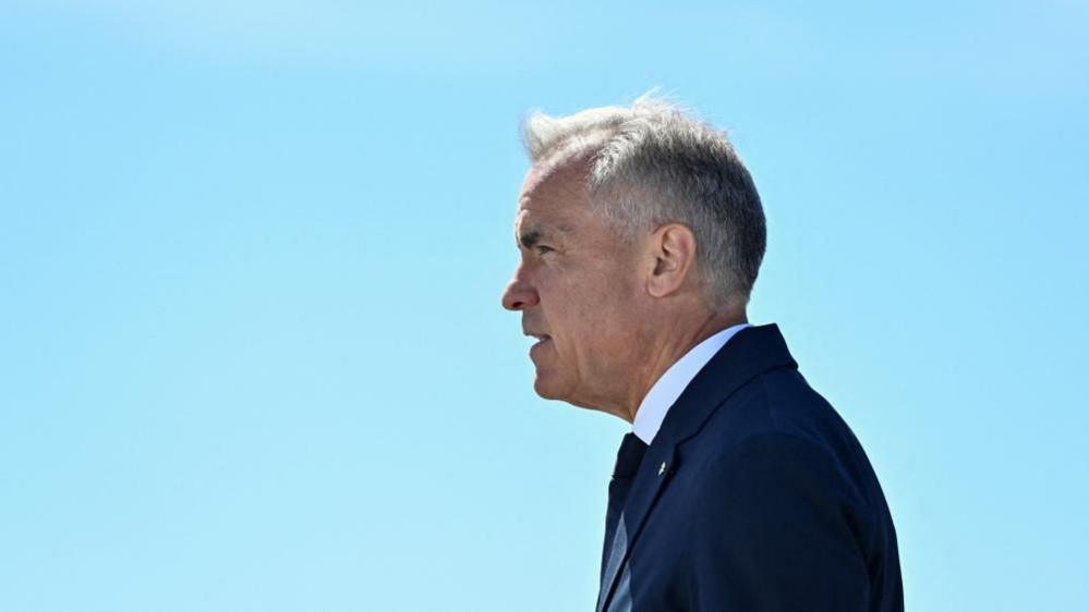 Mark Carney stands in profile against a clear blue sky. His hair is short and grey, and he wears a navy suit and tie and a white short. 