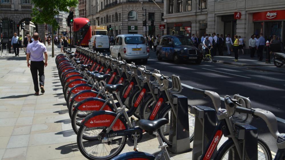 A long row of Santander Cycles bicycles are parked at a docking station, with a red wheel cover on the back wheel displaying the Santander logo. They are parked on a pavement along the side of a road, with taxis, buses and other traffic passing in front of them on the right. A few pedestrians are walking past, and some shops are visible on the street in the background.