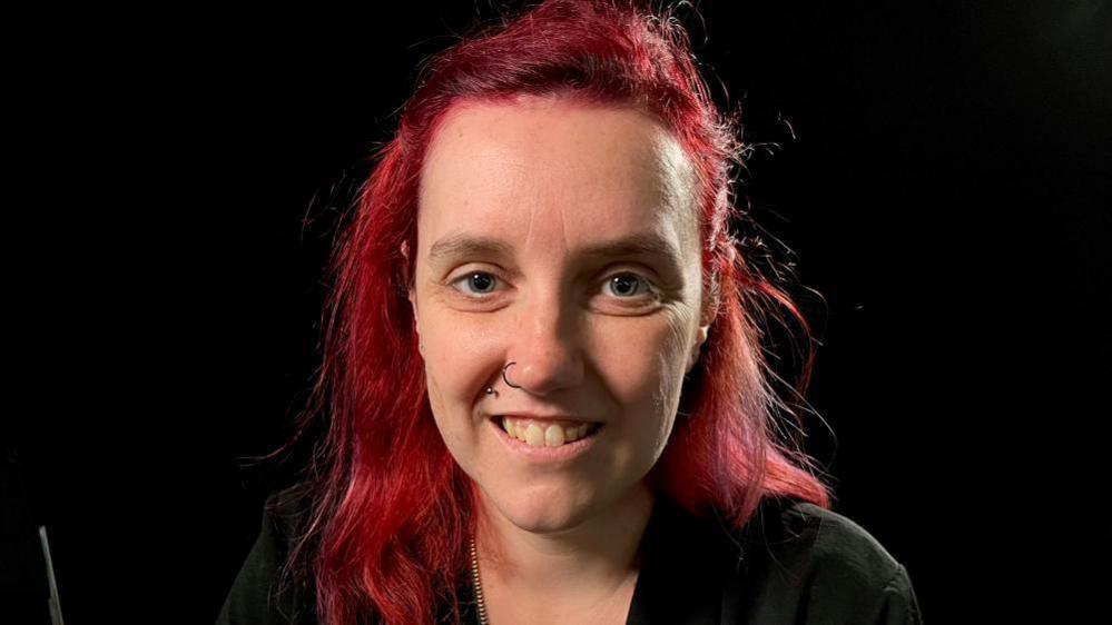 A picture of a woman with died red hair and a nose ring. she is smiling at the camera  and there is just a black back drop. 