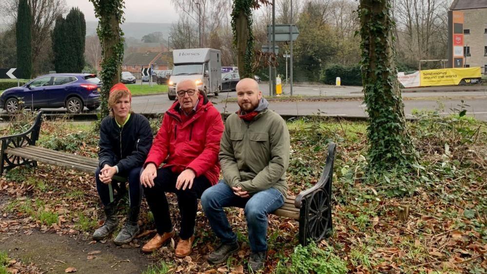 Three people with a solemn expression on their faces sit on a broken bench next to a busy road with cars and vans behind them