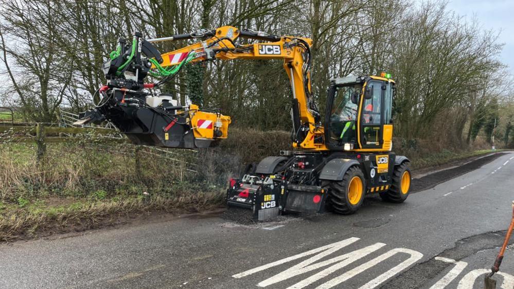 Yellow JCB digger with basket on the end of an extendable arm.  It is on a road with the word "SLOW" on one side. The road is lined by trees.