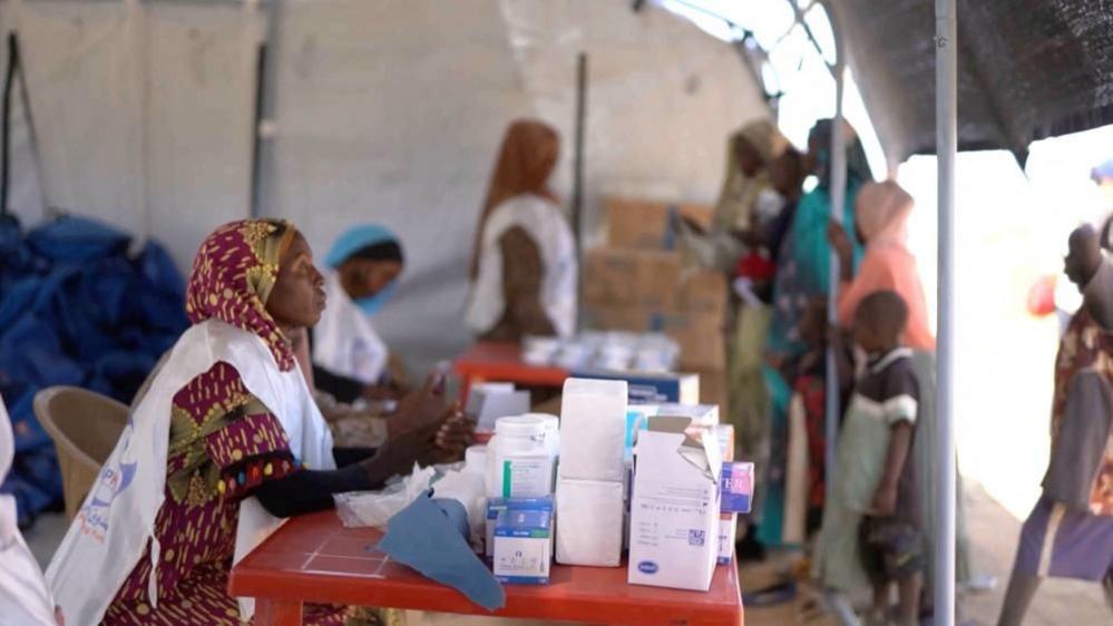 A medic waits in a makeshift clinic as displaced Sudanese gather. There is a large pile of white boxes in front of her.