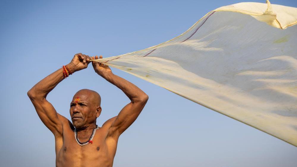 Image shows a Hindu devotee drying his cloth after taking a holy dip at Sangam, the confluence of the Ganges and Yamuna rivers with the mythical, invisible Saraswati river, during the "Maha Kumbh Mela", or the Great Pitcher Festival, in Prayagraj, India, 28 January, 2025