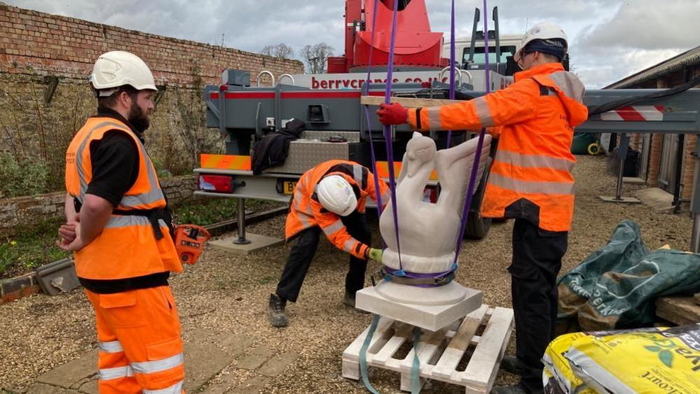 The stone scultpure being secured to the crane's winch by two operatives 