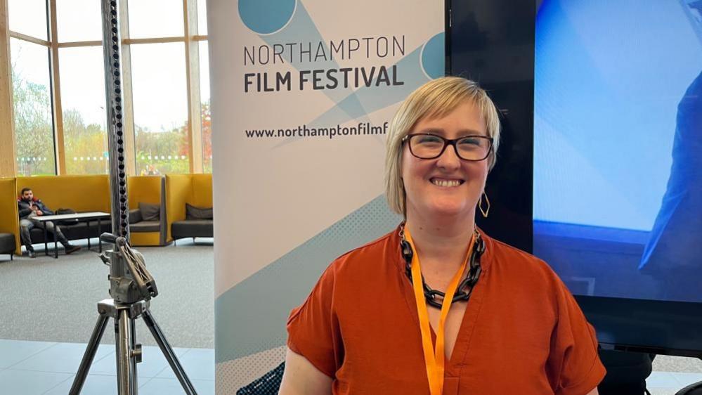 Becky with medium-length blond hair wearing a dark orange top and light orange lanyard as well as large brown glasses.  A board behind her says "Northampton Film Festival" and long windows are visible to the left