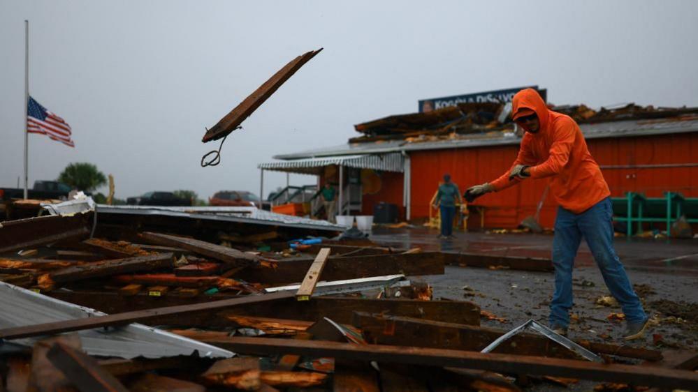 A man in a bright orange jacket tosses a plank of wood onto a pile during a hurricane clean up. An American flag at half mast is behind him, as is on orange building with a damaged roof and a sign on it. 