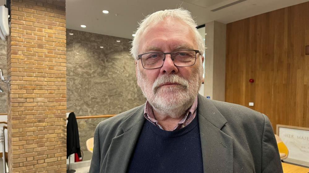 Steve Morphew is wearing a red checked shirt, a blue jersey and brown/grey suit jacket. He has white hair and a white beard and moustache, and is wearing black-rimmed glasses. He is standing in the lobby outside the Norfolk County Council chamber, with walls clad with brick, stone and wood panelling.