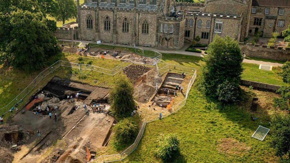 A drone shot of archaeologists digging in fenced off trenches on a grass slope, with Auckland Palace rising behind it.
