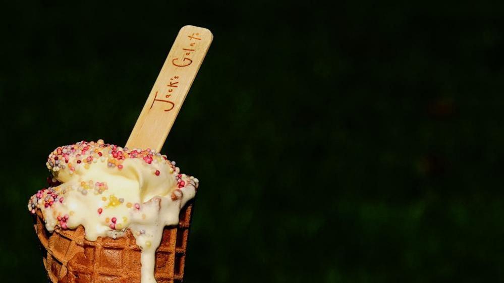 Ice cream, dotted with multi-coloured 100s and 1,000s, spilling over a waffle cone. A wooden spoon branded "Jack's Gelato" is sticking out of the top.