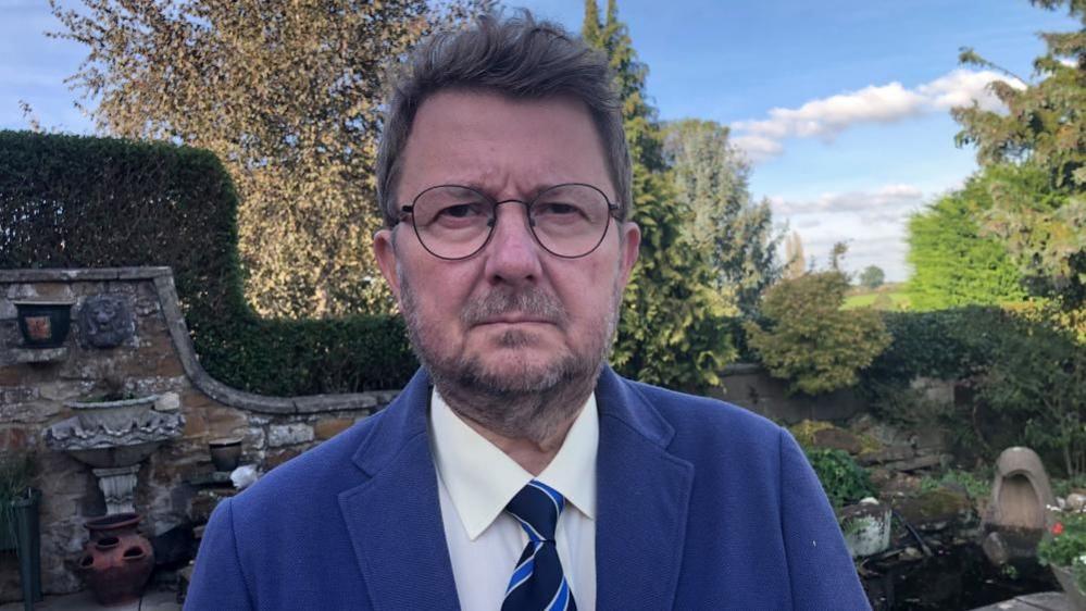 Sam Dobbs with short brown hair and beard wearing a blue jacket and tie in a garden