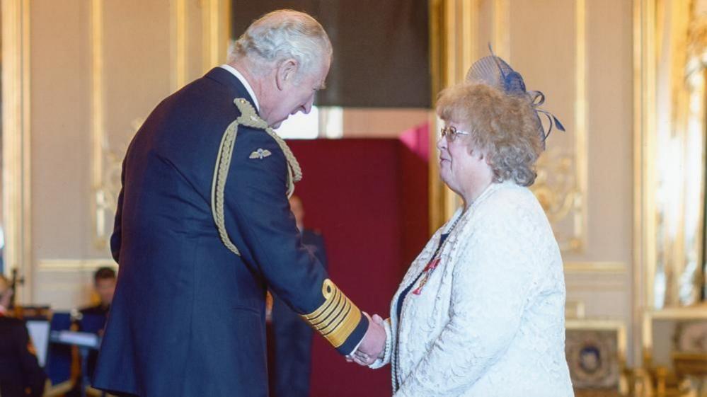 Diana Staines, in a white jacket, with a blue fascinator, being presented her MBE by King Charles III. He is wearing a blue military jacket featuring gold embroidered bands on the cuff and a lanyard on his shoulder.