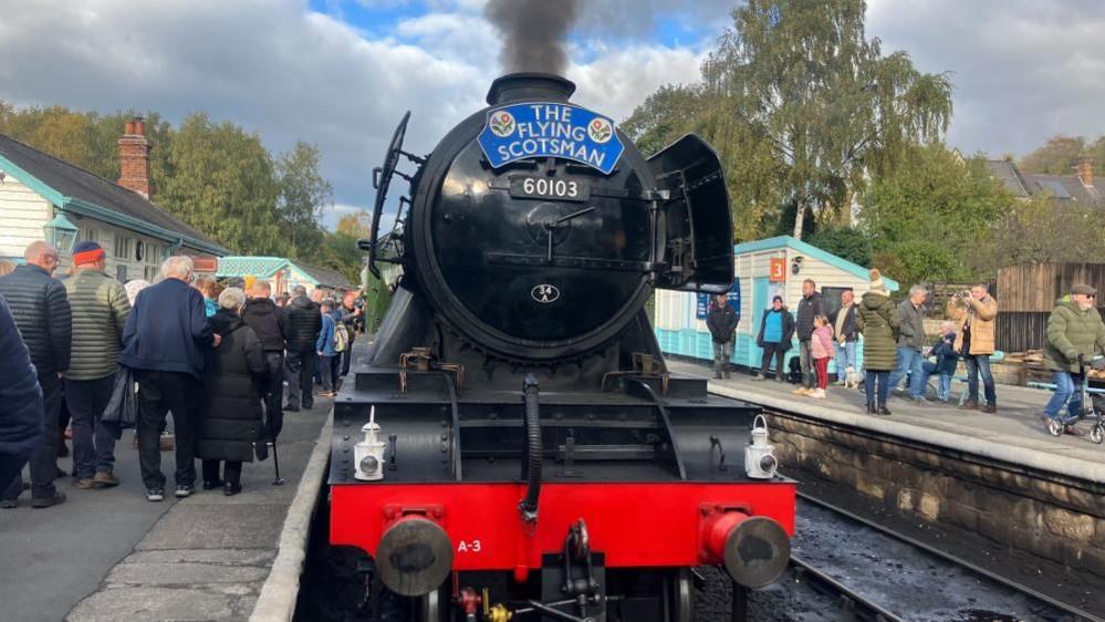 The front a a black stream train can be seen on railway tracks with crowds of people gathered on either side holding their phones and cameras up to take pictures. The engine appears to be 'in steam' with black smoke coming from the chimney. The blue nameplate on the engine reads 'The Flying Scotsman' and the number 60103 can be seen in gold print.