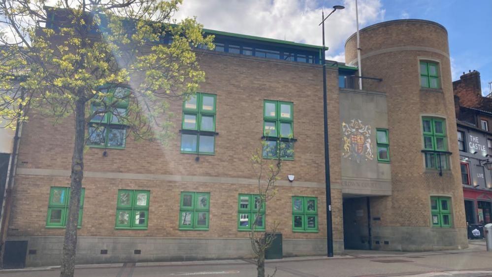 Luton Crown Court: A brown-brick building with green-framed windows. The words "CROWN COURT" written above the entrance on a grey wall below a multi-coloured crest.