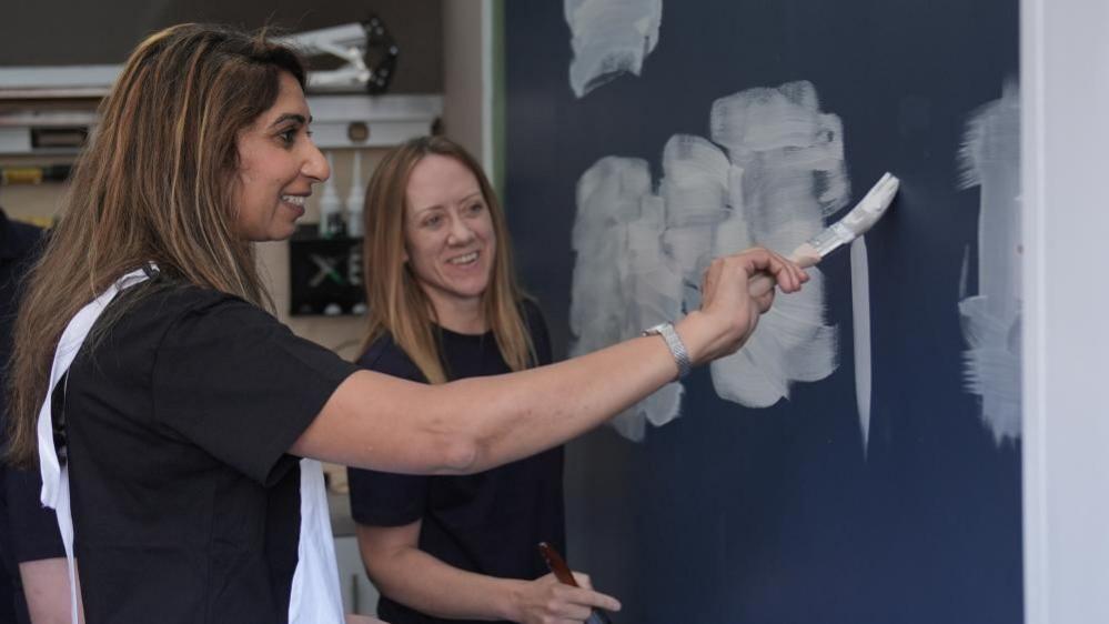 Suella Braverman is wearing a dark-coloured top and white dungarees. She is holding a paintbrush and painting white paint on to a blue wall. She is being observed by a woman in the background, who is wearing a black top.