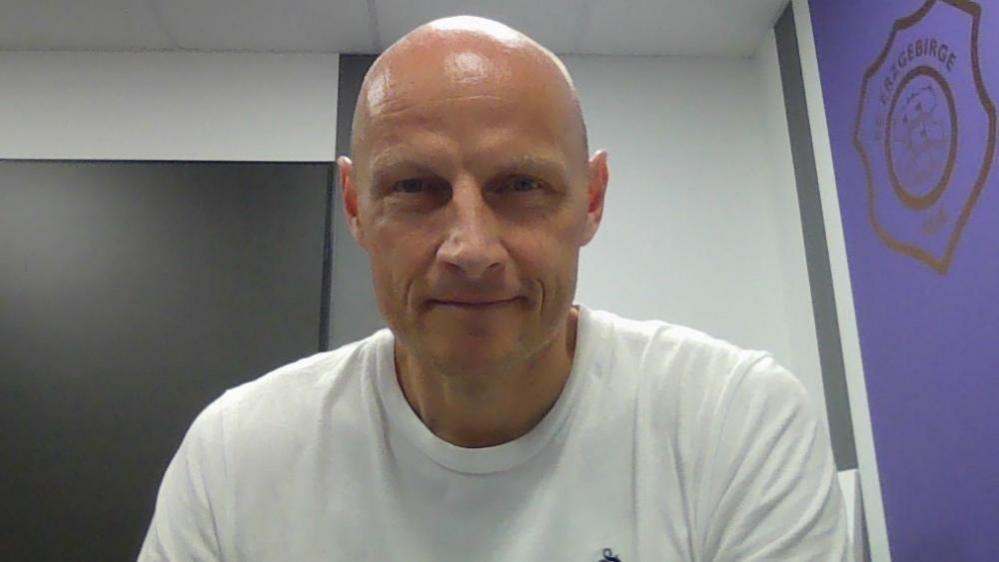 A bald man smiles into the camera. He is wearing a white t-shirt. On his left, there is a purple wall with the badge of German football club Erzgebirge Aue.