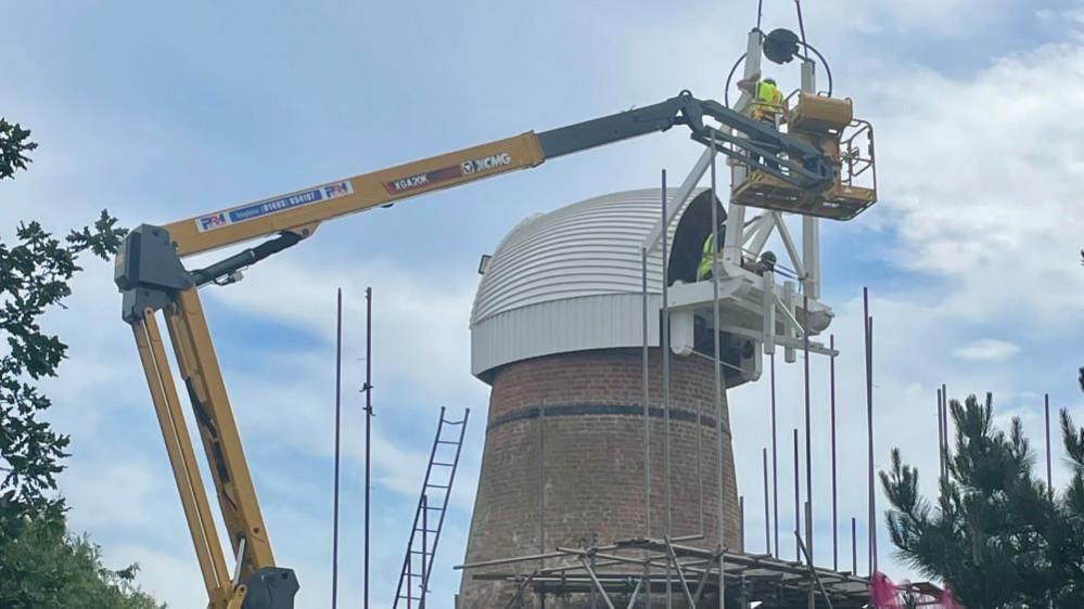 A man in a cherry picker works on one of the stocks of the Toft Monks Wind Pump, which features a white weather-boarded cap, with tail fan structure to the right of the image.