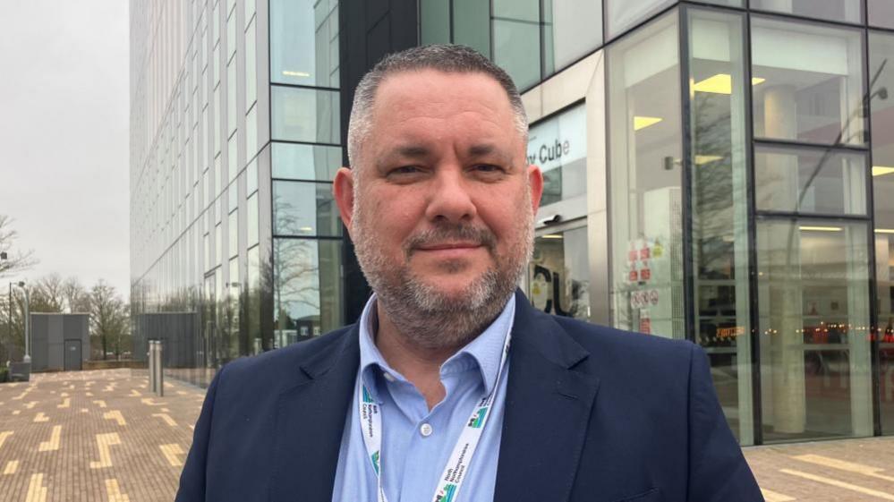 Jason Smithers with short dark hair and beard wearing a blue jacket and light blue short with a white and green council lanyard. He is standing in front of the Corby Cube, a modern four-storey glass and steel building.