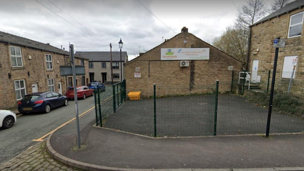 A Google Streetview screengrab of Cornerhouse Day Nursery in Lees. It is a brick building with a paved area outside and a fence running around the perimeter.