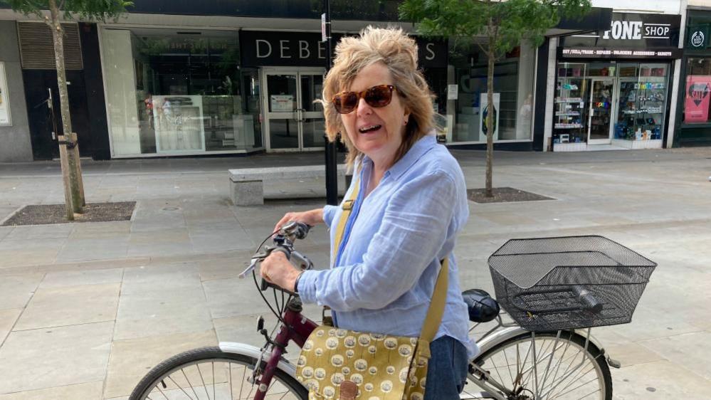 Kay Orton outside the former Debenhams in the centre of Bedford, pushing a bike, with a bag across her body, wearing a blue top 